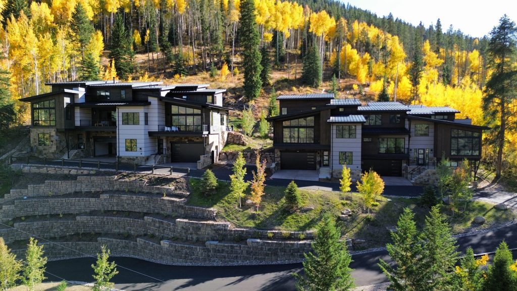 Modern duplexes built by H&C Construction in a Colorado mountain setting, featuring tiered landscaping, wood and stone siding, and large windows surrounded by fall foliage